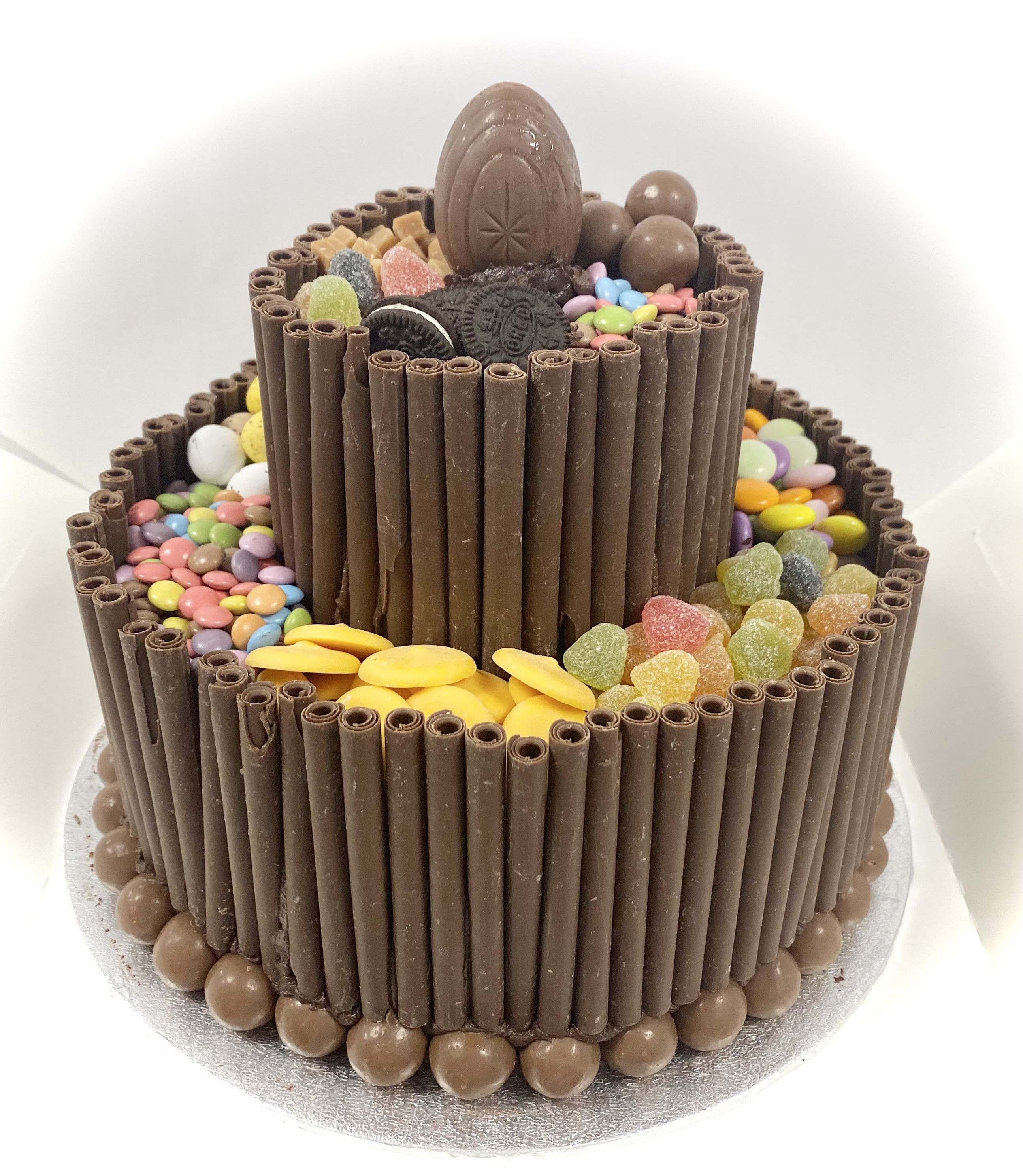 A two-tiered Chocolate Blackout Cake decorated with vertical chocolate sticks on the sides. The layers are topped with colorful candies, chocolate frosting, gummy hearts, and a large chocolate egg at the top. The cake sits on a white surface.